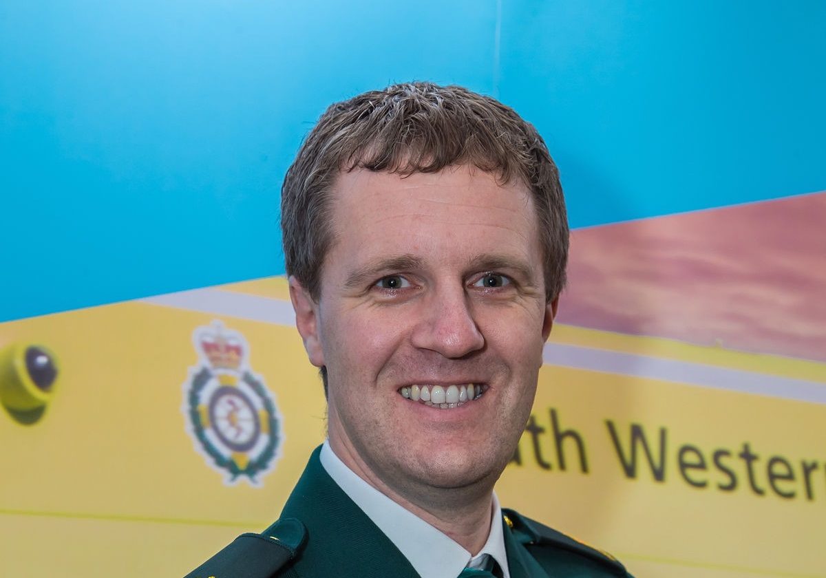 photo of man with short cropped hair wearing a green suit posing in front of a backdrop image of an ambulance